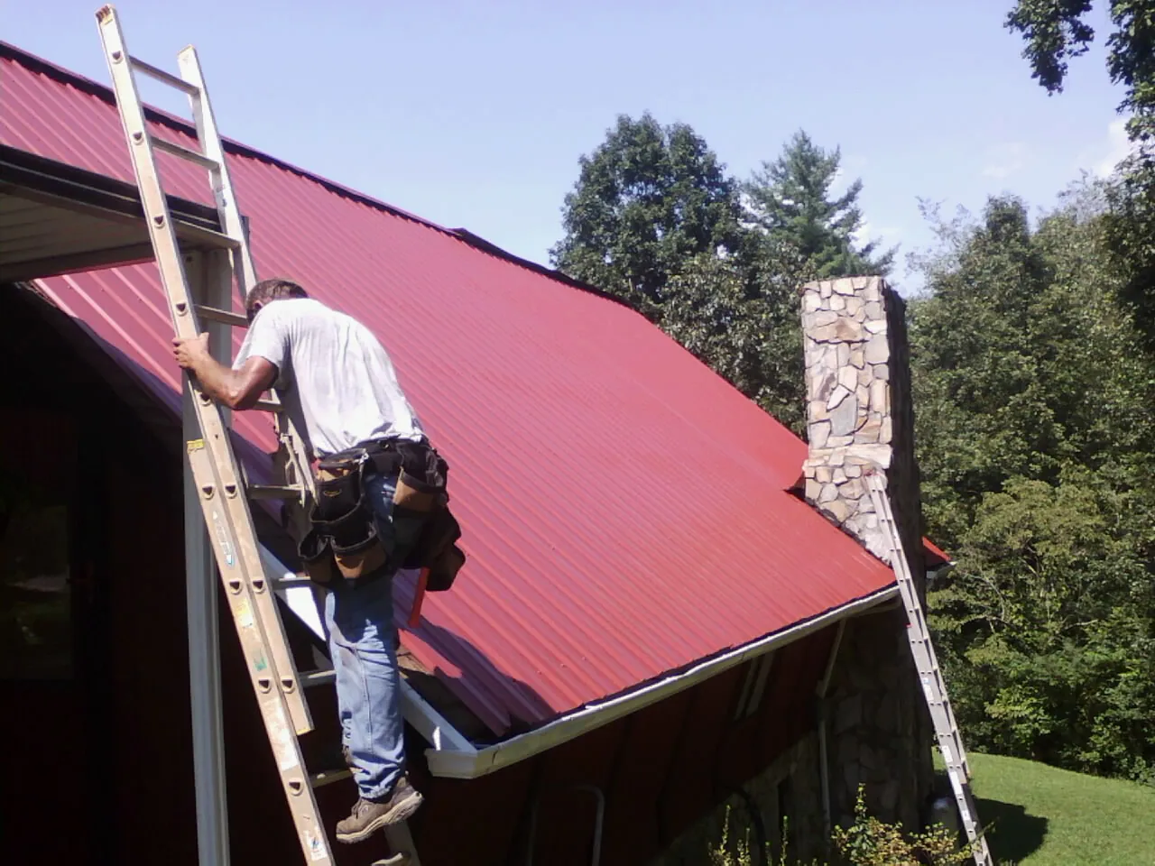 Roofer on a ladder beside a red metal roof and stone chimney — Soffit & Fascia Repair by Roofing Brothers in Walnut Creek